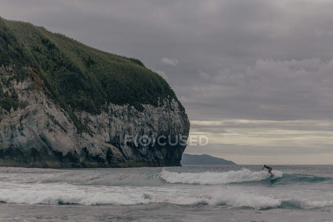 Pessoa surfar em ondas altas em mar agitado perto de rocha enorme no fundo do céu sombrio cinza — Fotografia de Stock