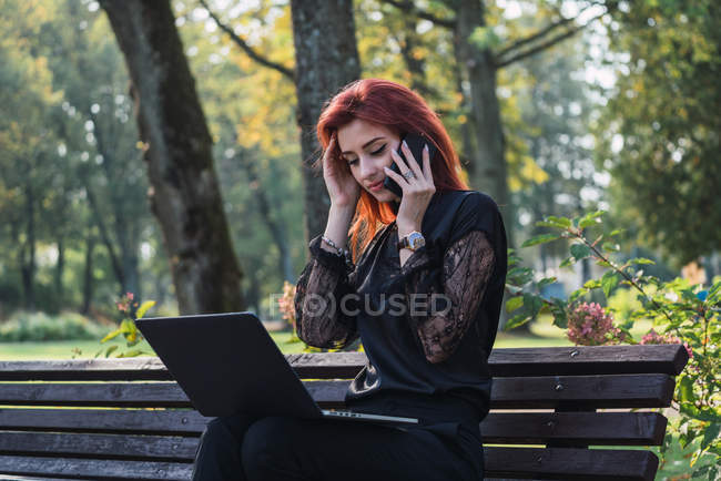 Mulher usando laptop e smartphone no banco no parque — Fotografia de Stock