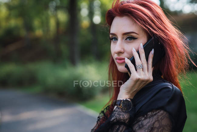 Mujer joven con el pelo de jengibre hablando en el teléfono inteligente en el parque soleado - foto de stock