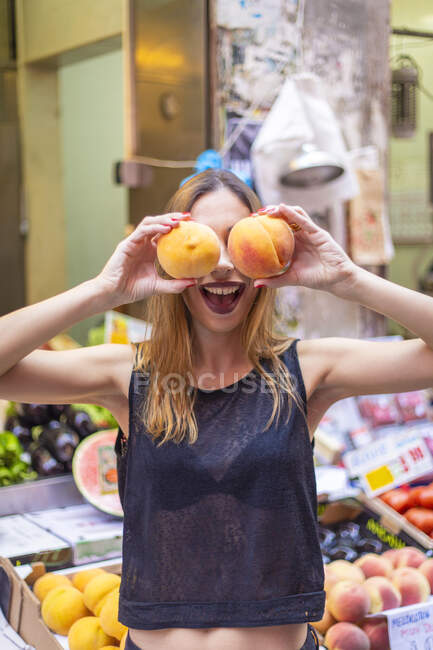 Lovely young female in stylish outfit keeping ripe peaches near eyes while standing near market stall — Stock Photo