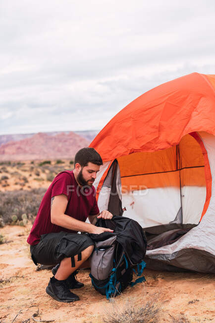 Vista laterale di bel ragazzo in abito casual imballaggio zaino mentre seduto a terra vicino tenda sulla zona campeggio nel deserto — Foto stock