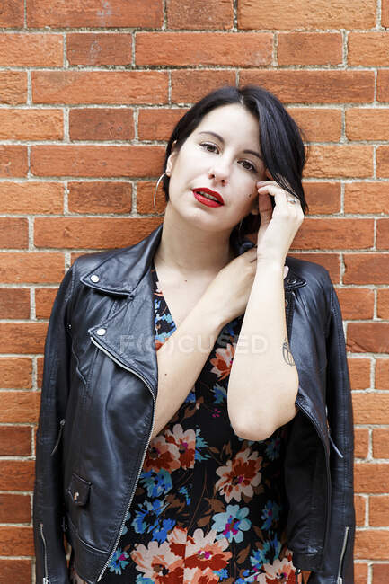 Young beautiful woman in leather jacket and dress with flower design standing near brick wall and looking at camera — Stock Photo