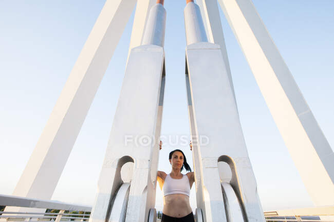 Adult fit woman in sportive top standing between pillars of contemporary steel bridge construction in sunlight looking at camera — Stock Photo