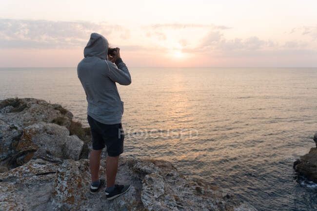 Seitenansicht eines nicht wiederzuerkennenden Mannes, der auf einer rauen Klippe steht und den herrlichen Sonnenuntergang über dem Meer in Tyulenovo, Bulgarien, fotografiert — Stockfoto