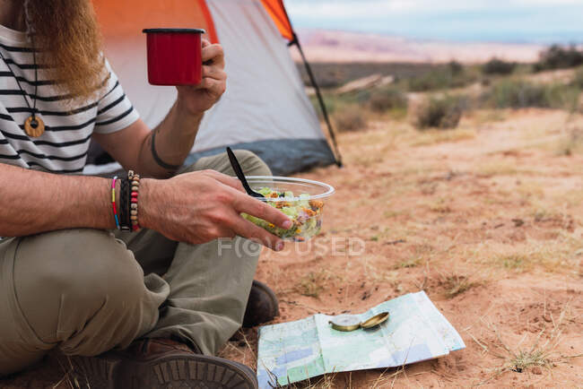Crop man eating salad and enjoying hot drink while sitting on sandy ground near map and compass during camping in desert — Stock Photo