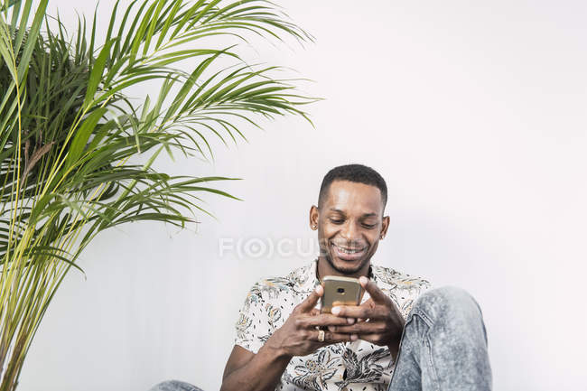 Sorrindo homem negro usando smartphone contra parede branca com planta verde — Fotografia de Stock
