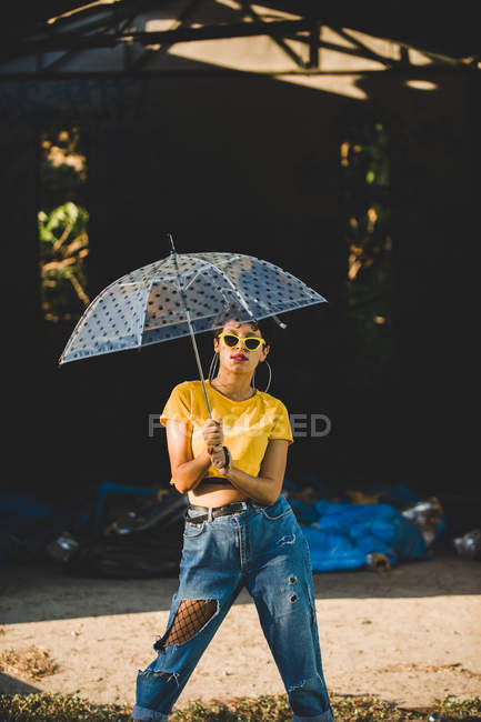 Charmante junge Frau in stylischem Outfit mit durchsichtigem Regenschirm, während sie an sonnigen Tagen auf der Straße steht — Stockfoto