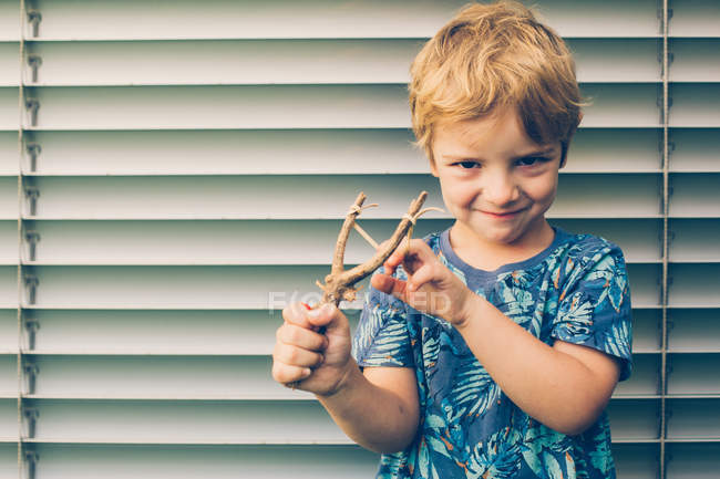 Блондинка маленький хлопчик у футболці грає з рогатками проти віконниць — стокове фото