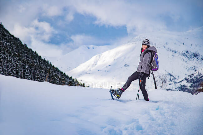 Vista lateral da fêmea em roupas quentes levantando a perna com esqui enquanto está em pé na encosta da montanha nevada — Fotografia de Stock
