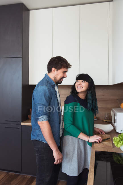 Vista laterale di eccitato giovane signora dando pomodoro tagliato al fidanzato allegro durante la cottura in cucina elegante insieme — Foto stock