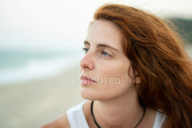 Hermosa joven con cabello de jengibre mirando hacia otro lado mientras está de pie sobre un fondo borroso de playa y mar en Tyulenovo, Bulgaria - foto de stock