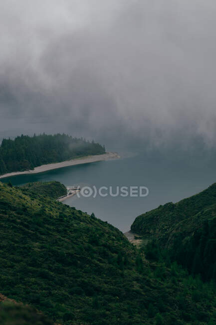 From above view of beautiful pure lake surrounded by mountains with green trees with thick fog above — Stock Photo