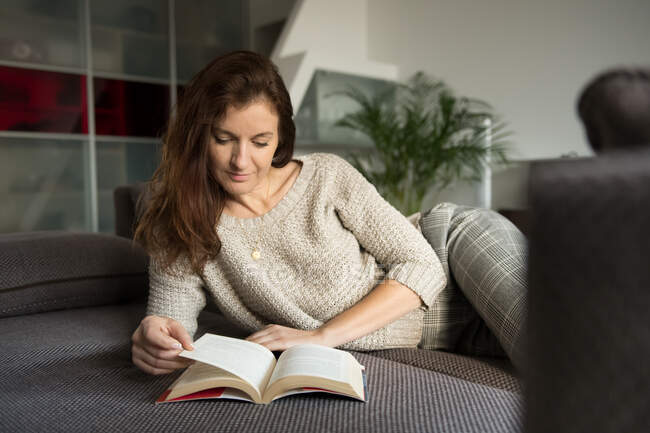 Lovely adult lady in elegant outfit smiling and looking away while lying on comfortable sofa with interesting book — Stock Photo