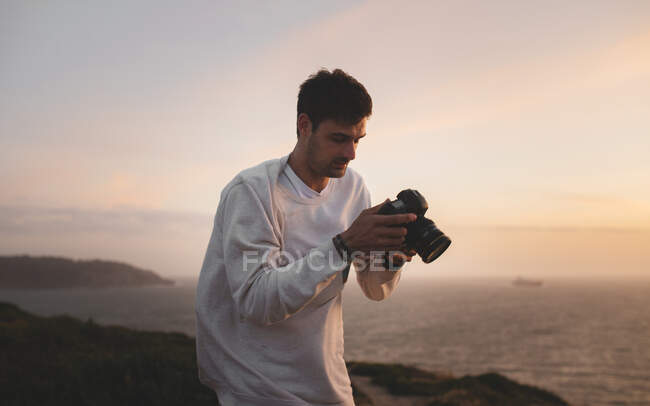 Наручний чоловік, що тримає фотокамеру і дивиться на фотографії, стоячи на скелі під час вітряного вечора у Сан - Франциско. — стокове фото