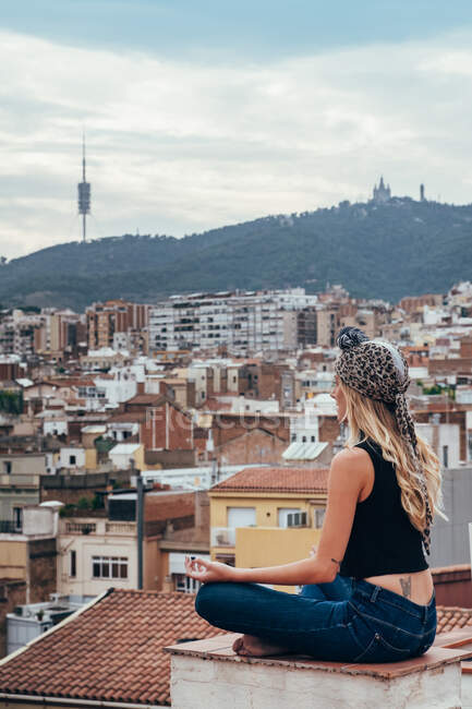 Вид збоку приваблива блондинка в голові, сидячи на даху в медитаційному положенні і дивлячись на мальовничий міський пейзаж — стокове фото
