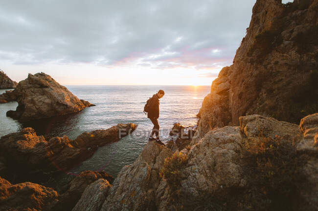 Vista laterale di una persona irriconoscibile in piedi su una ruvida roccia vicino alla calma acqua di mare durante il tramonto in Costa Brava, Spagna — Foto stock