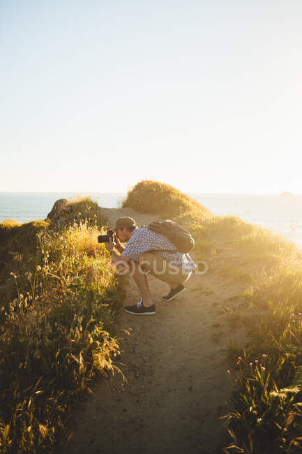 Vista lateral de un joven usando una cámara fotográfica profesional para tomar fotos de increíble naturaleza durante la puesta de sol en la playa en California - foto de stock