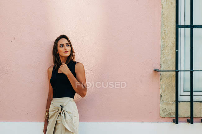 Charmante femme brune en tenue tendance penchée sur le mur sur la rue et regardant ailleurs — Photo de stock