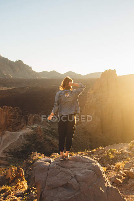 Vista posterior de la joven hembra de pie sobre piedra en el hermoso desierto y admirando la vista de la increíble puesta de sol en la isla de Tenerife en España - foto de stock