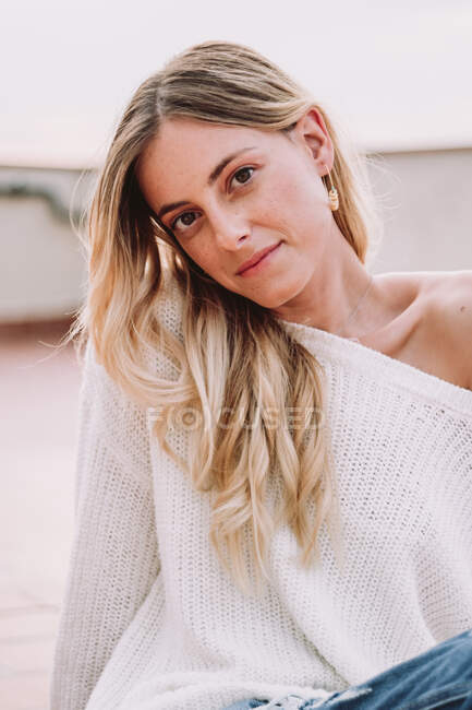 Junge hübsche Frau mit blonden Haaren in weißem Pullover sitzt draußen bei Tageslicht und blickt in die Kamera — Stockfoto