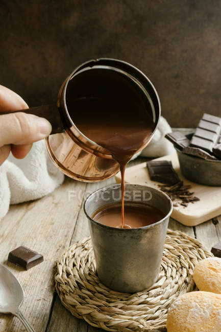 Human hand pouring hot chocolate from cooper pot into metal cup on wooden table — Stock Photo