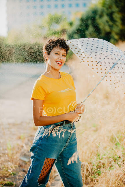 Junge Frau mit Regenschirm in stylischem Outfit lächelt und schaut in die Kamera, während sie unter Tropfen sprühenden Wassers läuft — Stockfoto
