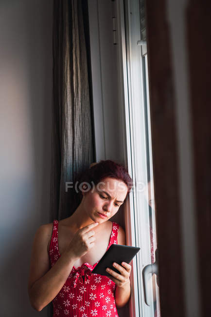 Mujer joven apoyada en la pared junto a la ventana con teléfono móvil - foto de stock