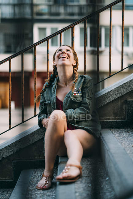 Сміється рудоволоса дівчина з косами, сидячи на сходах міста — стокове фото