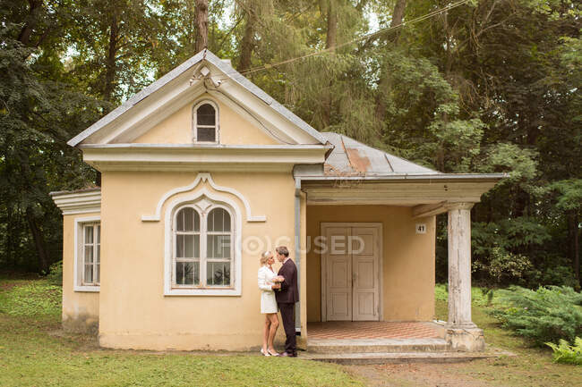Vista lateral da noiva elegante e noivo abraçando perto de pequena casa velha no jardim verde — Fotografia de Stock