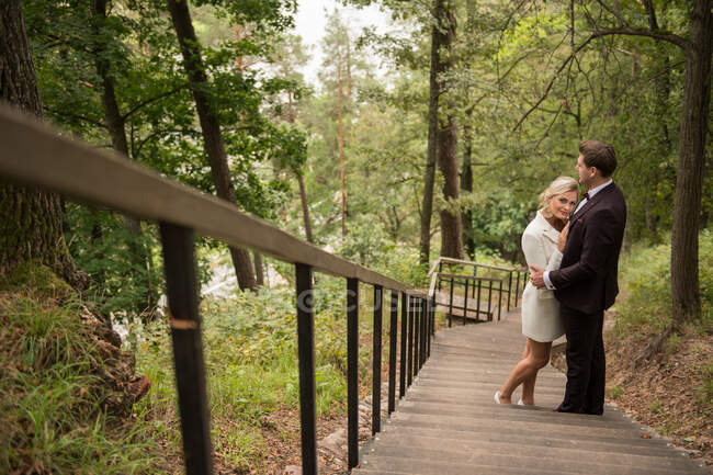 From above shot of embracing adult bride and groom standing on wooden walkway in green woods — Stock Photo