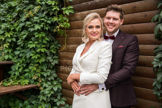 Elegante erwachsene Braut und Bräutigam umarmen sich, während sie in Anzügen vor einer Holzwand stehen und in die Kamera lächeln — Stockfoto