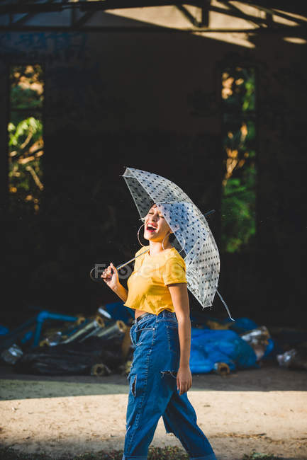 Очаровательная молодая женщина в стильном наряде смеется и держит прозрачный зонтик, стоя на улице в солнечный день — стоковое фото