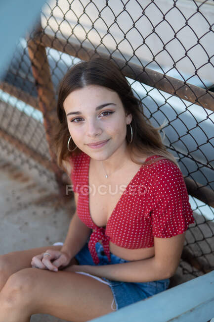 Desde arriba de la encantadora joven en traje de verano sentado en la valla de alambre en la calle mirando a la cámara - foto de stock