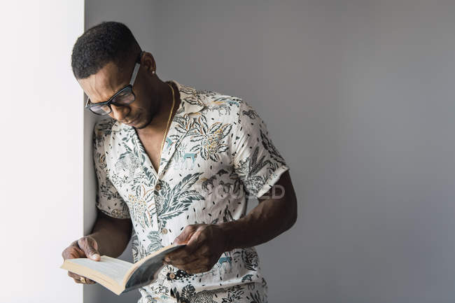 Uomo nero casual in occhiali appoggiato sulla parete bianca vicino alla finestra e libro di lettura — Foto stock