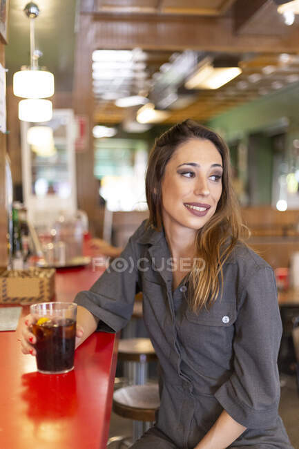 Beautiful young lady with glass of cold beverage smiling and looking away while sitting near counter in stylish cafe — Stock Photo