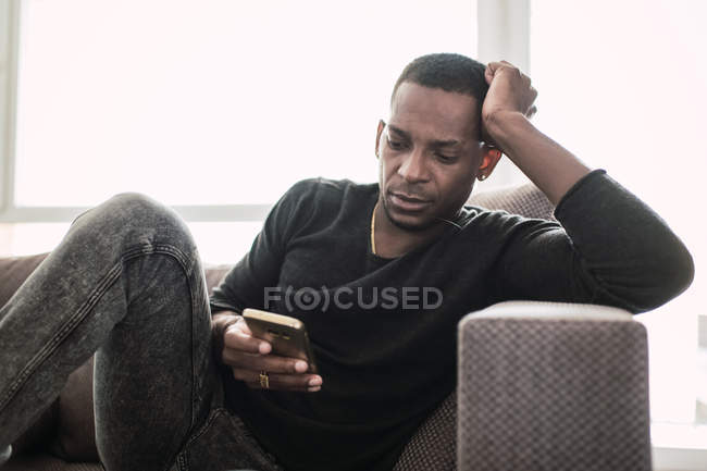 Lässiger afrikanisch-amerikanischer Mann sitzt zu Hause auf der Couch und surft mit angelehntem Smartphone — Stockfoto
