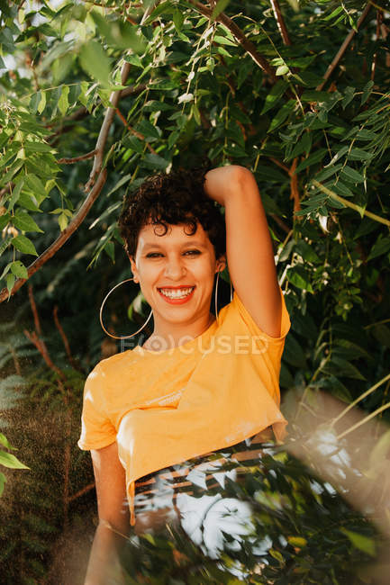 Портрет усміхненої брюнетки з коротким волоссям, що стоїть у зеленій рослинності з сонячним світлом — стокове фото