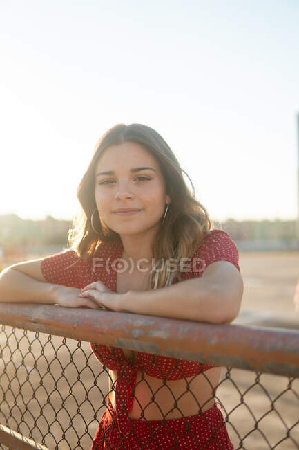 Jeune brune heureuse en tenue décontractée appuyée sur une clôture métallique et regardant la caméra au soleil — Photo de stock