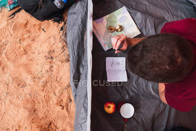 De arriba plano de chico joven acostado en la tienda y dibujo en cuaderno de bocetos mientras acampa en el desierto - foto de stock