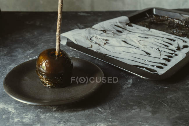 Black caramel apple on stick on plate on grey tabletop — Stock Photo
