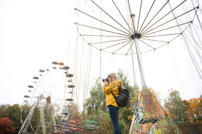 Vista posteriore di donna bionda con macchina fotografica che scatta foto di parco divertimenti desolato con attrazioni — Foto stock