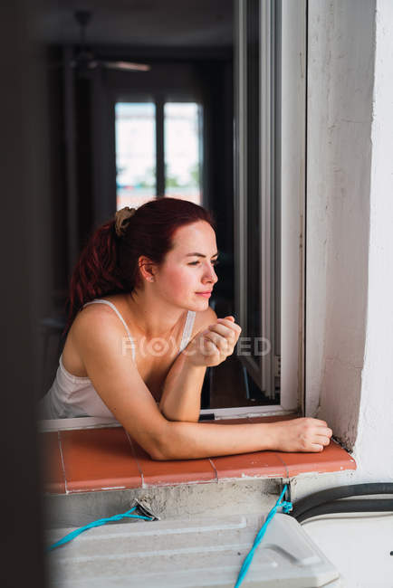 Jovem mulher de pé perto da janela aberta e olhando para a rua à luz do dia — Fotografia de Stock