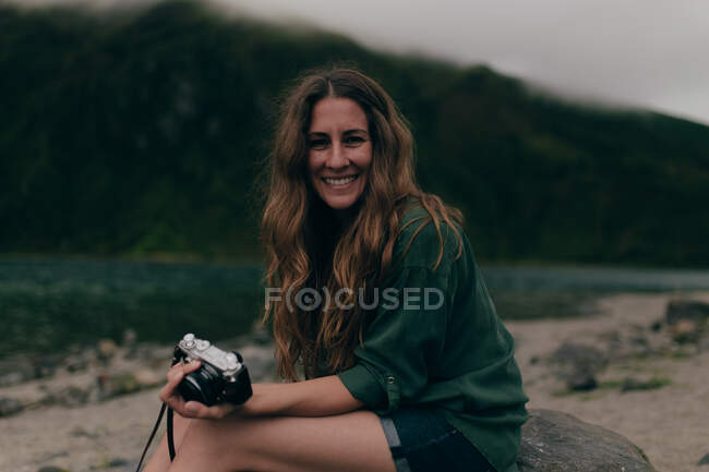 Vue latérale de belle jeune femme aux cheveux bouclés en chemise verte assise sur un rocher près du lac et regardant la caméra sur fond d'arbres — Photo de stock