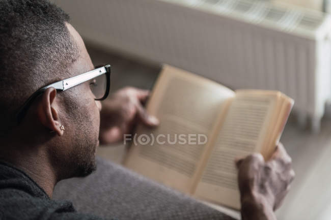 Close-up de homem afro-americano em óculos livro de leitura enquanto relaxa no sofá em casa — Fotografia de Stock