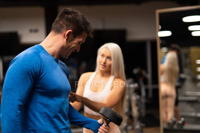 Femme aidant l'homme à faire de l'exercice avec haltère dans la salle de gym — Photo de stock
