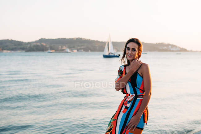 Содержательная женщина в длинном платье, стоящая на берегу на закате против моря — стоковое фото