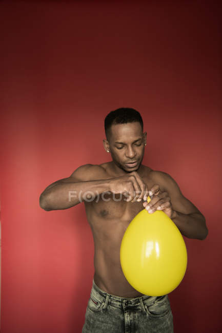 Muscular shirtless black man in jeans blowing bright yellow balloon on red background — Stock Photo