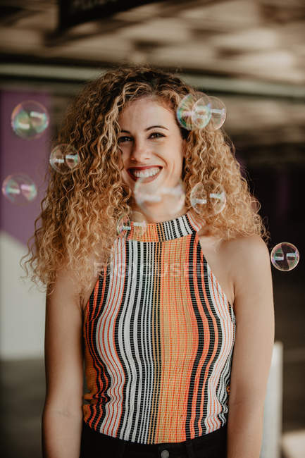 Playful blond curly woman standing among soap bubbles — Stock Photo
