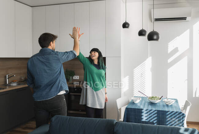 Alegre joven hombre y mujer dando cinco de altura el uno al otro, mientras que de pie en la cocina moderna juntos - foto de stock