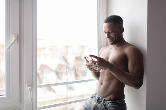 Hombre negro musculoso sin camisa usando el teléfono móvil mientras está de pie contra la ventana a la luz del día - foto de stock